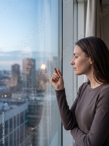 Woman alone with a wedding ring, lost in thought, gazing out the window at a city skyline
