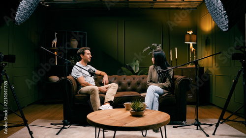 Man and woman producing a digital podcast, discussing topics into microphones during a video recording session in a professional studio