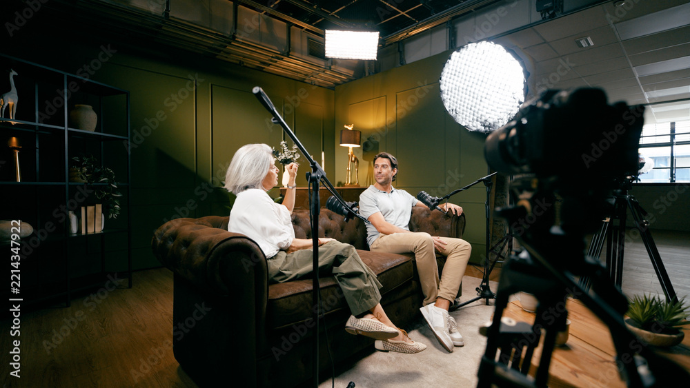 Fototapeta premium Two diverse podcasters sitting on a sofa in a studio setting, creating engaging audio and video content
