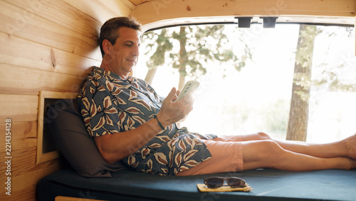 Man enjoying a comfortable vacation, lying on a bed inside a customized camper van and browsing his mobile phone