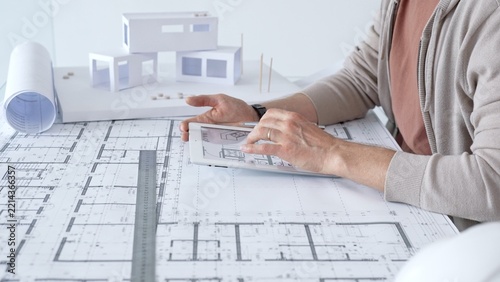 Architect working on a new building design using a tablet with digital blueprint plans. Also showing paper blueprints. A ruler. And a scale model of a house on the desk