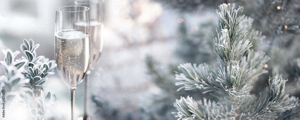 Fototapeta premium Two glasses of champagne with frozen branches in a snowy winter landscape. Horizontal background with fetsive bokeh for christmas and new year greetings.