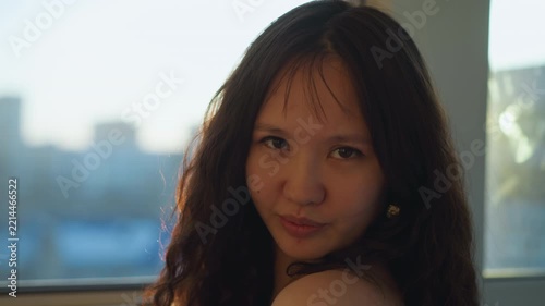 Woman with long wavy hair adjusts her head and looks out window to urban cityscape. Her loose curls catch soft daylight streaming through glass as woman stands near edge of viewable space