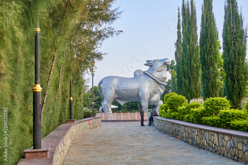 A large silver-colored statue of Nandi, the sacred bull in Hindu mythology, stands prominently at the end of a paved walkway. The sculpture features ornamental neck decorations and is surrounded by