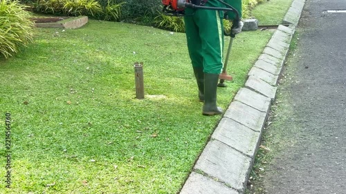 Worker mowing tall grass with electric or petrol lawn trimmer in city park or backyard. Gardening care tools and equipment. Process of lawn trimming with hand mower