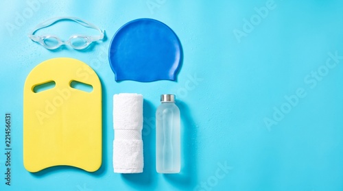 Flat Lay of Swimming Equipment Including Yellow Kickboard Blue Cap Goggles Towel and Water Bottle on Blue Background with Copy Space