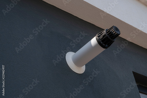 Exterior vent pipe on a wall. A white plastic exhaust pipe with a black louvered cap protrudes from a dark gray textured stucco wall near a roofline. Useful for HVAC, plumbing, and building.
