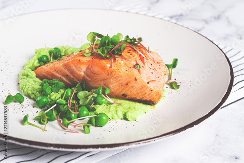 Grilled salmon filet served on a bed of green pea puree with fresh microgreens and peas, presented on a white plate