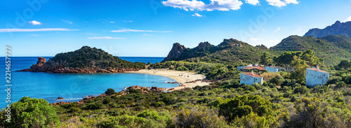 Rocky coastline in Sardinia, Italy, featuring striking rock formations and sand beach with lush greenery against a backdrop of clear blue sea. Rugged landscape provides a serene and natural escape