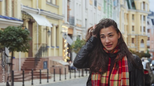 Cheerful brunette girl in colorful scarf laughing on cozy city street with historical buildings. Joyful woman enjoying autumn day in town