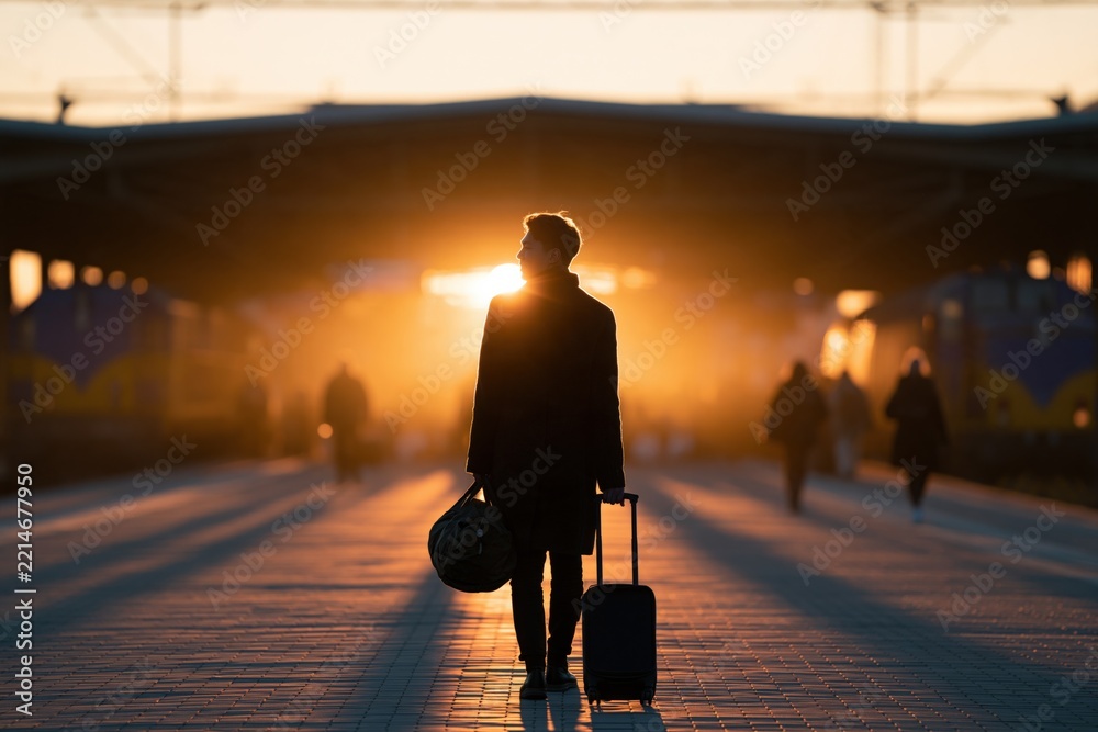 Fototapeta premium Silhouetted traveler with suitcase walking on a train station platform at sunrise for travel background and banner design