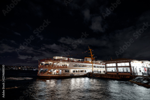 Bright Istanbul ferry moored at a waterfront terminal at night, reflecting across the Bosphorus under a deep blue sky.