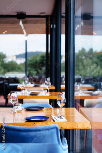 A modern restaurant interior featuring a wooden dining table set with plates, wine glasses and blue chairs, creating an inviting atmosphere for fine dining, socializing, and relaxed meals with a view.