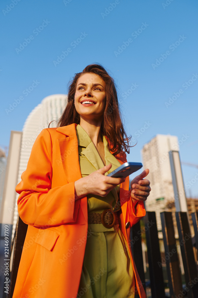 Fototapeta premium Smiling young woman in orange coat holds smartphone with both hands, texting outdoors against city skyline. Concept of messaging, digital connection and happy mobile communication.
