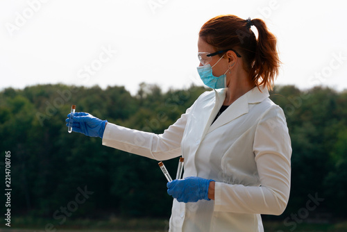 pneumonic plague outbreak scientist holding test tube sample, focused female epidemiologist in lab coat, face mask and blue gloves examining sample looking away from camera, forest and sky