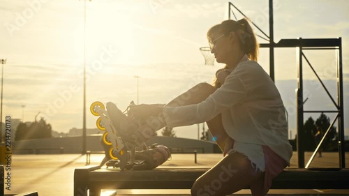 Young girl ties inline skates on bench at sunset. Blurry background shows active men playing basketball game on outdoor sports court during warm golden hour.