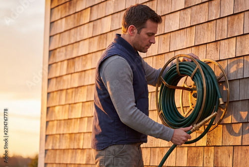 Man Coiling Green Garden Hose Onto Wall Mounted Reel at Golden Hour