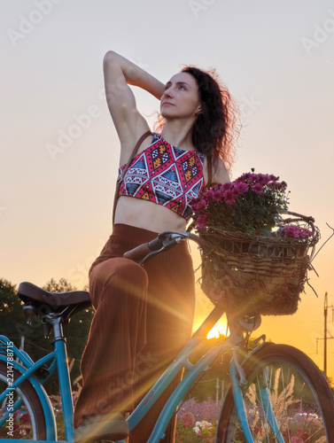Carefree attractive woman with long curly hair enjoying a beautiful summer evening ride on a classic blue bike with a flower basket, bathed in warm sunset backlighting outdoors.