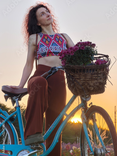 Young woman with dark curly hair posing with a vintage blue bicycle holding a basket of purple flowers during a warm golden sunset light in a picturesque field outdoors.
