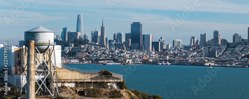 Historic water tower on Alcatraz Island stands above San Francisco Bay with downtown skyscrapers, Transamerica Pyramid, and urban waterfront hills visible under clear daylight skies.