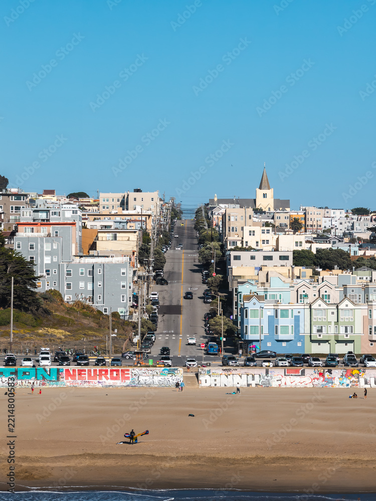 Fototapeta premium Ocean Beach seawall painted with graffiti fronts a sloping residential street with pastel houses and church steeple in San Francisco, California, viewed across wide sand.