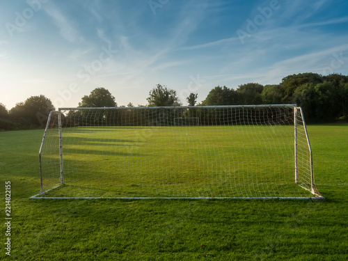 White soccer goal stands alone on green grass. Trees line the background under blue sky with wispy clouds. Scene peaceful and quiet, no people present. Ideal for sports, recreation