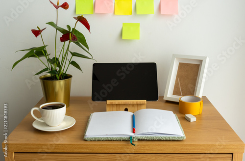Tablet with blank screen, open notebook and coffee on a wooden desk with colorful sticky notes and anthurium flowers. Concept of daily planning, productivity and working from home.