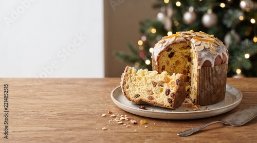 A delicious Panettone cake, adorned with icing and candied fruit, sits on a wooden table, with a blurred Christmas tree in the background, evoking a festive holiday atmosphere.