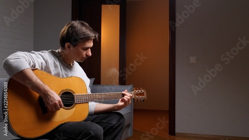 Young man sitting on a sofa in a modern home, strumming an acoustic guitar and practicing music for leisure, relaxed and focused on songwriting and skill building