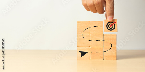 Hand completing a route of wooden cube blocks leading to a dartboard target. Business achievement, goal setting, objective planning, and growing business success concept.