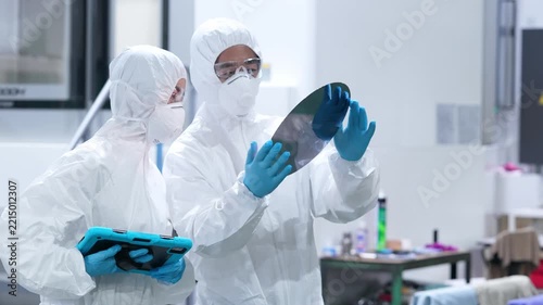 Male and female workers in protective suits inspect silicon wafers with tablets in a semiconductor factory.
