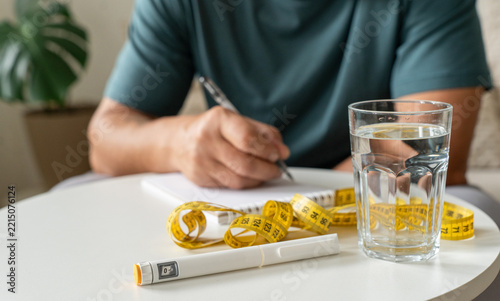 Man is taking notes at table where an injection pen, measuring tape, and glass of water are placed. He is tracking health metrics, weight loss progress, and metabolic control