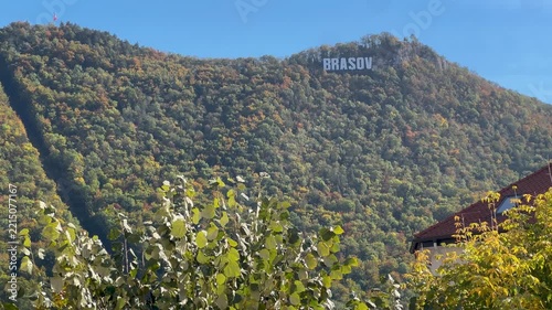 Scenic view of the famous Brasov sign atop Mount Tampa surrounded by colorful autumn forest under clear blue sky, capturing tranquil travel mood in Romania. 4K video.