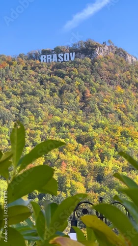 Scenic view of Mount Tampa with iconic Brasov sign surrounded by colorful autumn forest foliage and green foreground leaves under bright sunny blue sky in Romania. 4K vertical video.