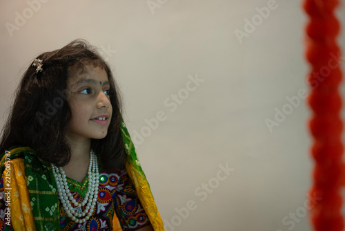 Portrait of a young Indian girl wearing traditional Gujarati ethnic clothing with a green patterned dupatta, colorful embroidered blouse, and layered pearl necklace.