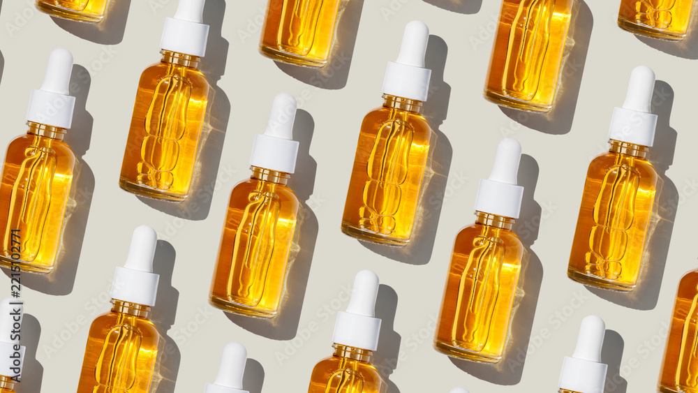 Fototapeta premium Minimal pattern glass dropper bottles filled with yellow golden face serum at sunlight on neutral fon. Minimal cosmetic product for skincare, anti-aging, and beauty concept, repeated layout, top view