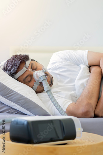 Man wearing a CPAP breathing mask sleeping on a bed with a CPAP machine