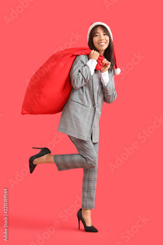 Young Asian businesswoman in Santa hat with bag of gifts on red background