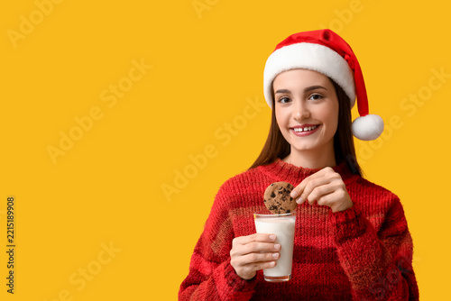 Young woman in Santa hat with milk and cookie on yellow background