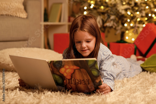 Cute happy little girl in pajamas with book reading Christmas story at night