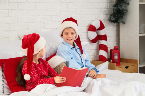 Cute little children with Santa hats reading book in bedroom on Christmas eve
