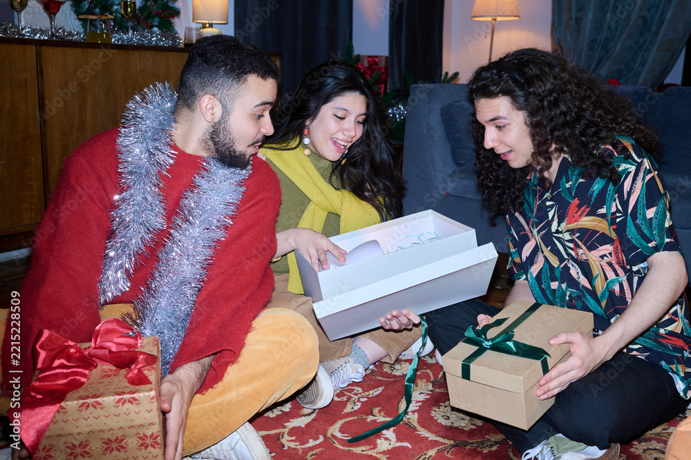 Fototapeta premium Multiethnic group of young adults opening gift boxes during Christmas celebration at home. Scene suitable for holiday advertising, festive retail campaigns, and seasonal promotion