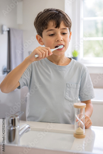 Child brushing teeth with sand timer in bright bathroom. Young boy focuses on brushing teeth with toothbrush while sand timer measures time. Concept of dental hygiene for kids and parenting tips.