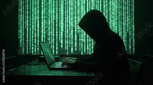 Anonymous hacker in hoodie typing on laptop with green binary code background in dark room.