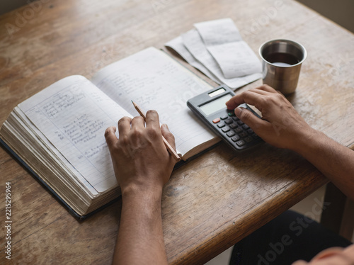Overhead view of hands writing in a household ledger and using a calculator at a kitchen table