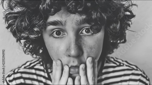 A young person with curly hair slowly lowers their hands from their face to reveal wide, startled eyes in a black and white.