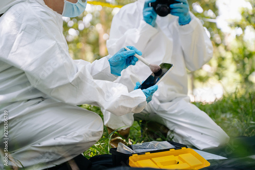 Forensic investigators in protective hazmat suits meticulously dust a smartphone for fingerprints and photograph evidence at a woodland crime scene
