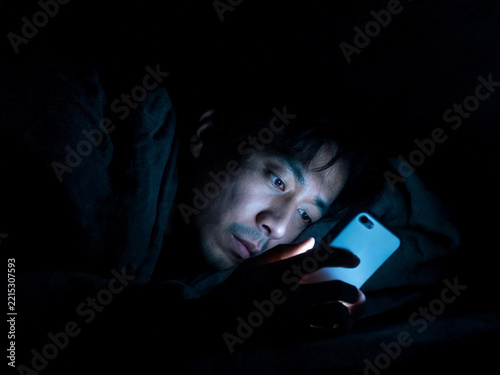 Tired Asian man lying in bed at night scrolling smartphone in the dark, blue light on face, insomnia and digital fatigue concept
