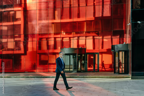Businessman walks outdoors past a modern red glass office building