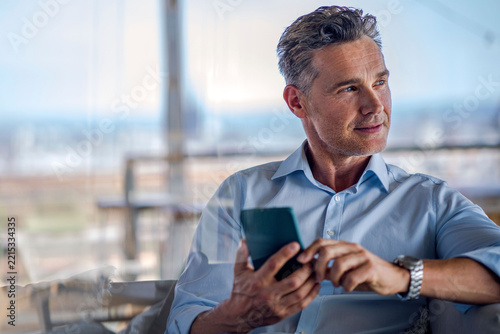Thoughtful businessman holding smartphone by window in office